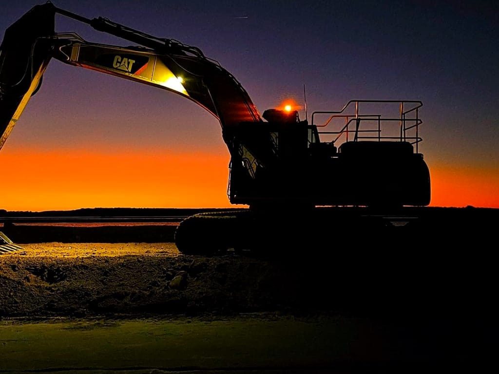 A CAT excavator standing in silhouette against a burnt orange dusk sky at the end of a shift