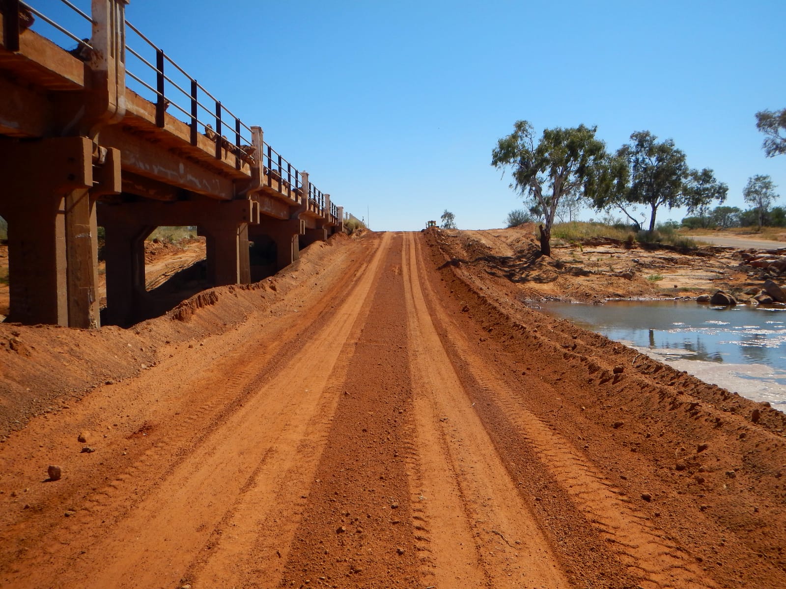 A freshly resheeted red gravel road running past a low concrete bridge over a Murchison river pool, graded flat with windrows either side